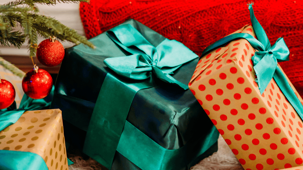 A close-up of festively wrapped gifts next to a Christmas tree, with a red knitted blanket in the background.