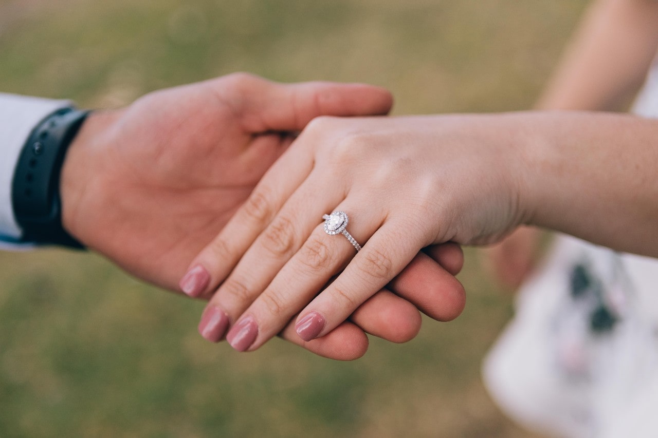 A close-up of a newly engaged couple holding hands, a delicate halo ring with an oval center stone on the bride-to-be’s finger. A close-up of a newly engaged couple holding hands, a delicate halo ring with an oval center stone on the bride-to-be’s finger.