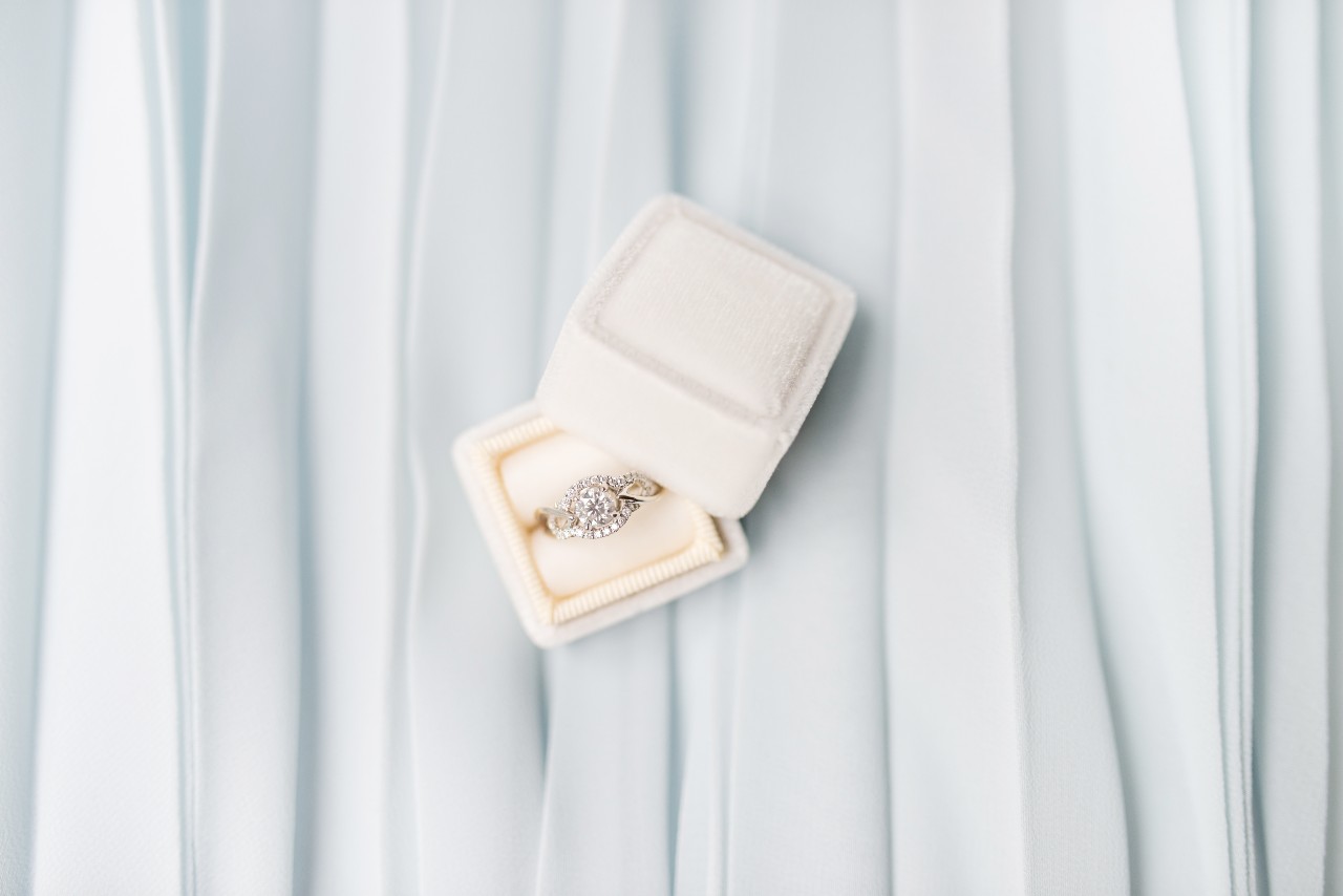 A close-up of a dazzling halo engagement ring displayed in a cream-colored box on a textured backdrop. A close-up of a dazzling halo engagement ring displayed in a cream-colored box on a textured backdrop.