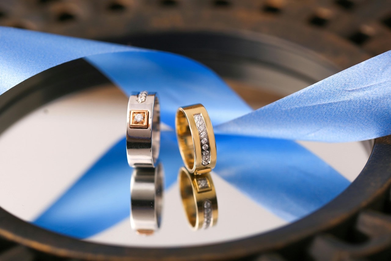 Two mixed metal and diamond wedding bands sitting on a mirror with a blue ribbon. Two mixed metal and diamond wedding bands sitting on a mirror with a blue ribbon.