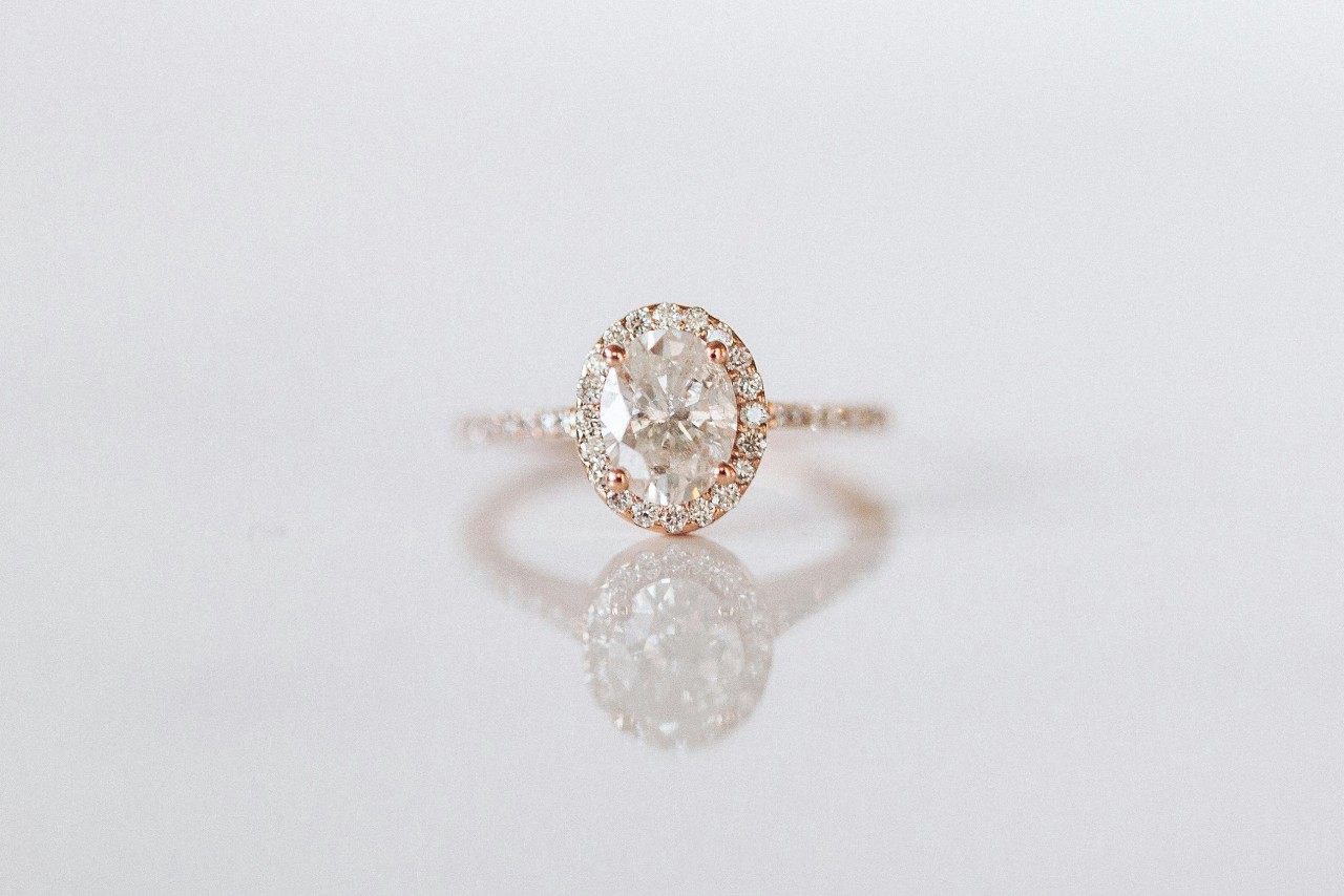 A close-up of a rose gold with diamonds around the band and a halo around the oval center stone diamond that is sitting on a reflective white background. A close-up of a rose gold with diamonds around the band and a halo around the oval center stone diamond that is sitting on a reflective white background.