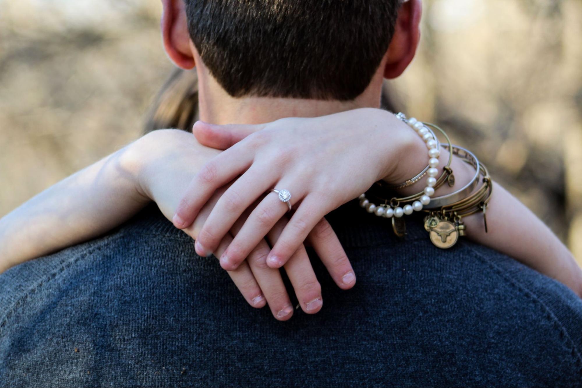 A close up of a woman's hand hugging a man, showcasing engagement ring, pearl bracelet, and stacked gold bangles.