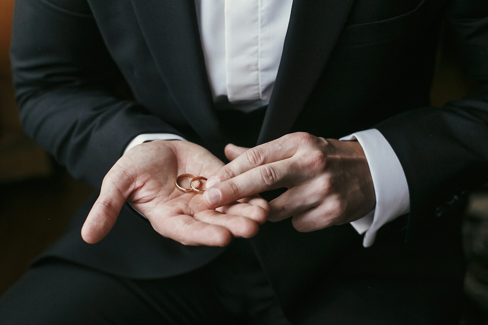 A close-up of a groom in a black suit holding two gold wedding rings in his palm.