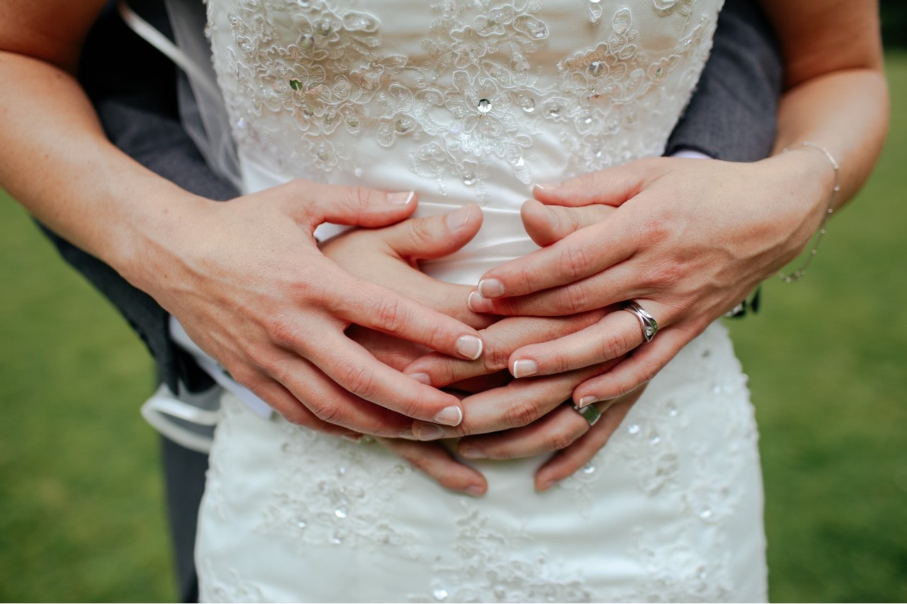 A close-up of a bride and groom's hands lovingly intertwined as he embraces her from behind.