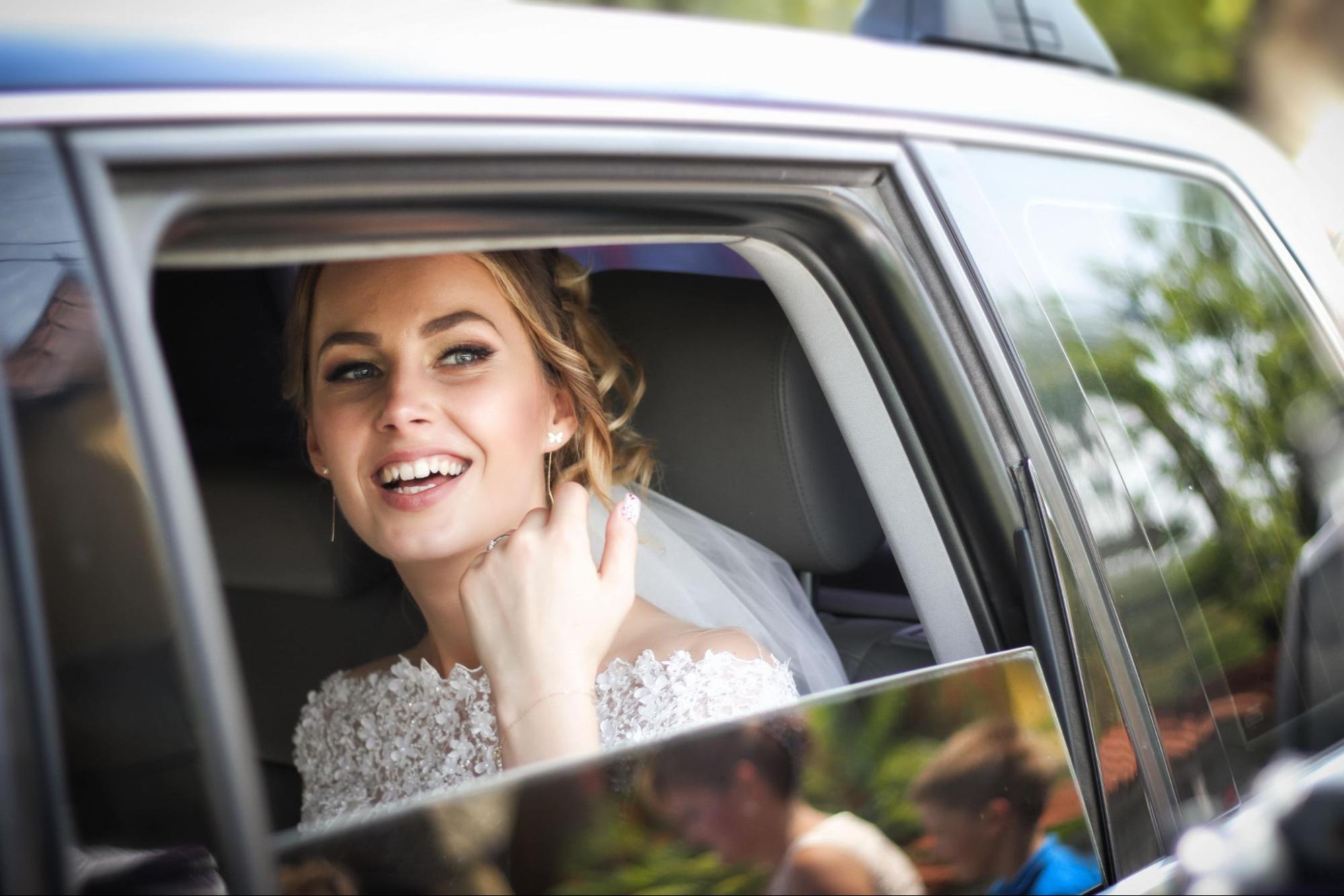 A smiling bride in a lace dress is seated inside a car, looking out of a partially open window.
