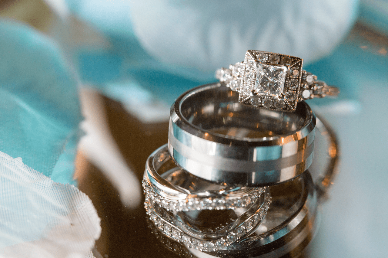Close-up of two white gold wedding bands and an ornate diamond engagement ring with a square setting on a reflective surface, surrounded by soft blue fabric. Close-up of two white gold wedding bands and an ornate diamond engagement ring with a square setting on a reflective surface, surrounded by soft blue fabric.