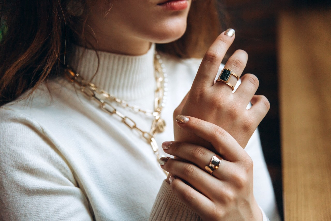A woman wearing a white turtleneck, adorned with yellow gold jewelry, including chain link and fashion rings. A woman wearing a white turtleneck, adorned with yellow gold jewelry, including chain link and fashion rings.