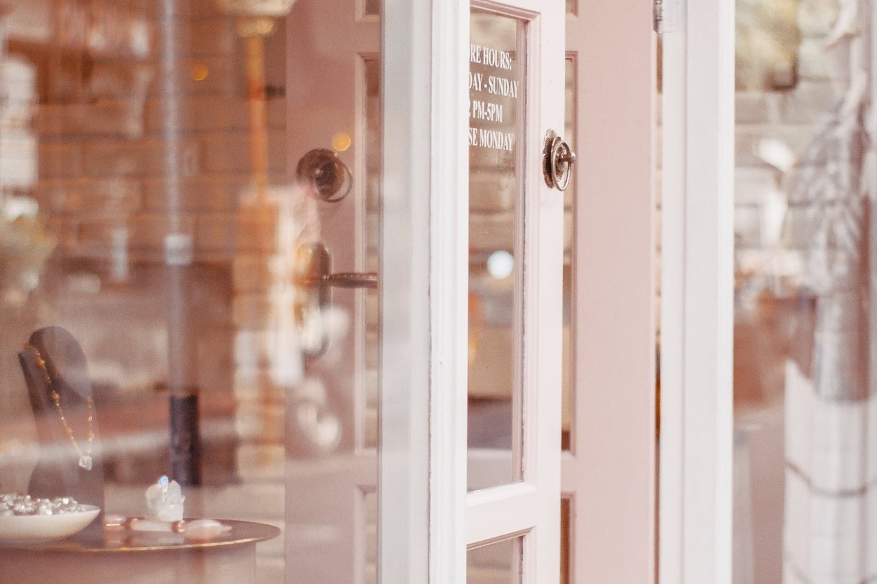 A soft pink door of a jewelry store is partially open, revealing necklaces and crystals inside. A soft pink door of a jewelry store is partially open, revealing necklaces and crystals inside.