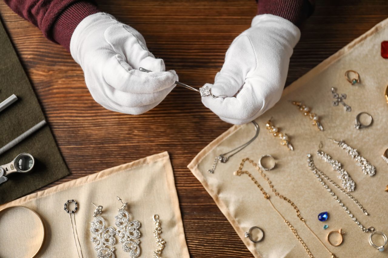 A jeweler wearing white gloves inspects a diamond ring using a tool, surrounded by various sparkling jewelry items on beige fabric. A jeweler wearing white gloves inspects a diamond ring using a tool, surrounded by various sparkling jewelry items on beige fabric.