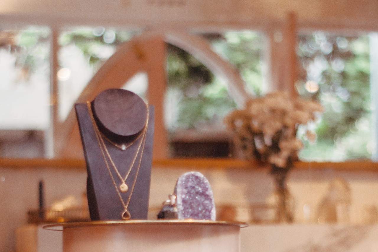 A close-up of a jewelry display with layered gold necklaces on a black stand inside a warmly lit boutique. A close-up of a jewelry display with layered gold necklaces on a black stand inside a warmly lit boutique.