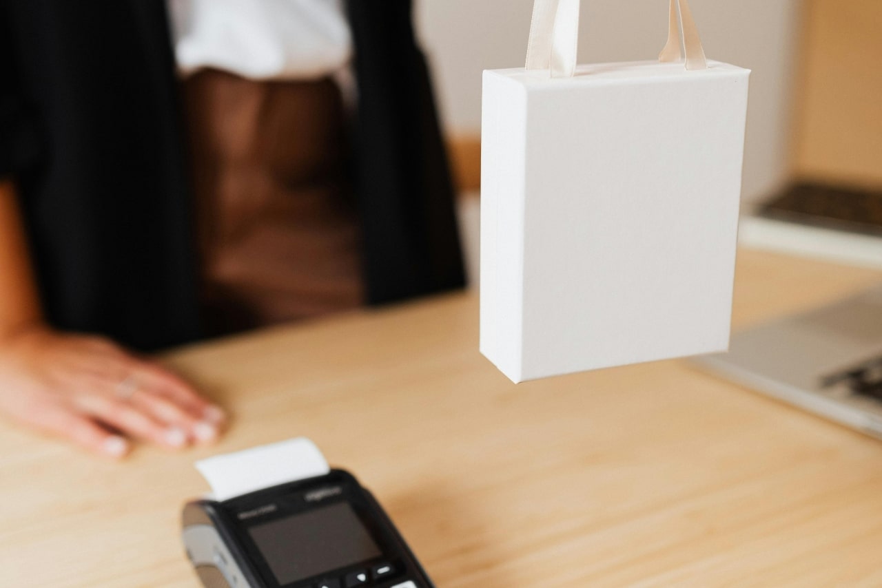 A close-up photo of a jeweler handing a white gift bag to a customer across a wooden counter. A close-up photo of a jeweler handing a white gift bag to a customer across a wooden counter.
