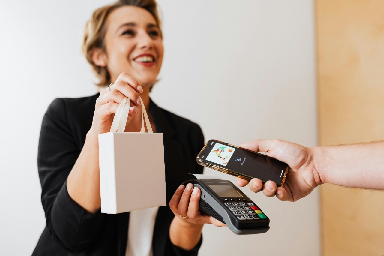 A smiling cashier holding a white shopping bag and card reader while customer pays with phone A smiling cashier holding a white shopping bag and card reader while customer pays with phone